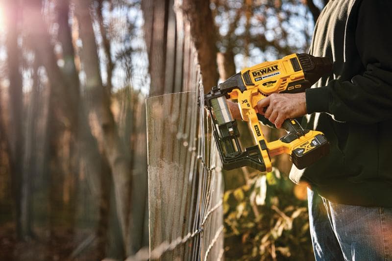 Una persona usando una grapadora inalámbrica para cercas DEWALT para asegurar una cerca de alambre al aire libre, con árboles y luz solar en el fondo.
