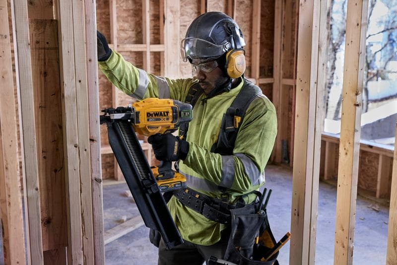 Un trabajador de la construcción con equipo de seguridad utiliza una clavadora DEWALT DCN920B para asegurar los marcos de madera dentro de un edificio en construcción. La cara del trabajador está desenfocada.