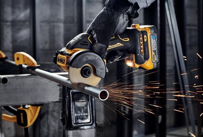 Una persona con guantes utiliza una amoladora angular inalámbrica brushless DEWALT para cortar una tubería de metal, generando chispas en un taller industrial.
