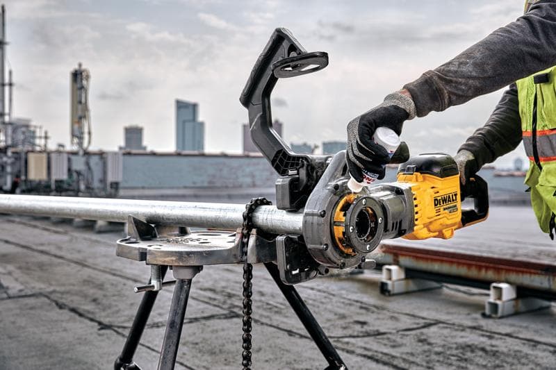 Una persona usando una herramienta eléctrica amarilla DEWALT DCE700X2 para roscar una tubería metálica montada en un soporte de trabajo, al aire libre en una azotea con edificios de la ciudad visibles al fondo. El usuario lleva guantes y un chaleco de seguridad.