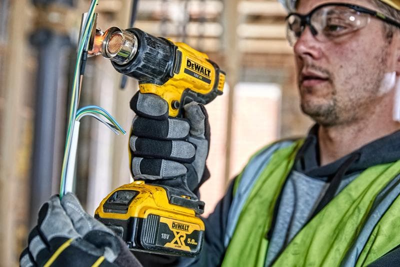 Una persona con guantes de trabajo y chaleco de seguridad usa una pistola de calor inalámbrica DEWALT DCE530N para aplicar calor a cables eléctricos en un sitio de construcción. El rostro de la persona está difuminado por privacidad.