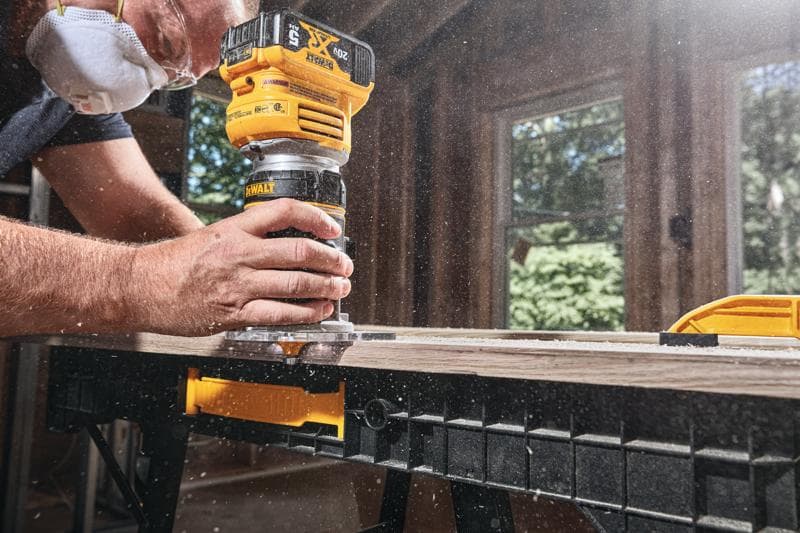 Primer plano de una persona usando una fresadora inalámbrica DEWALT para dar forma al borde de una tabla de madera en un banco de trabajo, con aserrín volando y la persona usando mascarilla protectora.