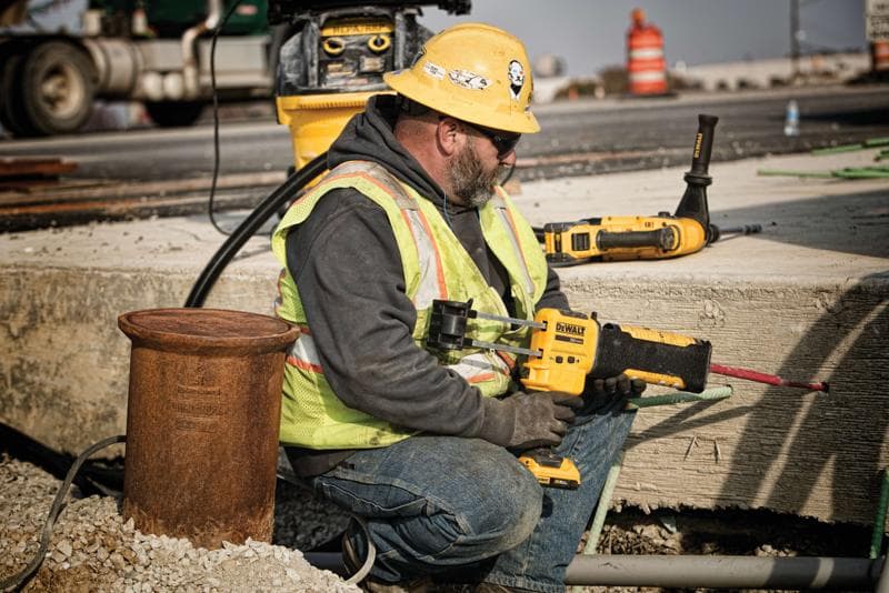 Un trabajador de la construcción con casco amarillo, chaleco de seguridad y guantes está usando una herramienta eléctrica DEWALT para perforar concreto en un sitio de construcción al aire libre. El rostro del trabajador está desenfocado por privacidad. Otras herramientas DEWALT y equipos de construcción se ven al fondo.