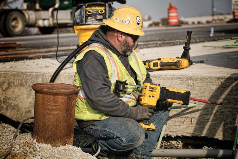 Un trabajador de la construcción con casco amarillo, chaleco de seguridad y guantes está usando una herramienta eléctrica DEWALT para perforar concreto en un sitio de construcción al aire libre. El rostro del trabajador está desenfocado por privacidad. Otras herramientas DEWALT y equipos de construcción se ven al fondo.